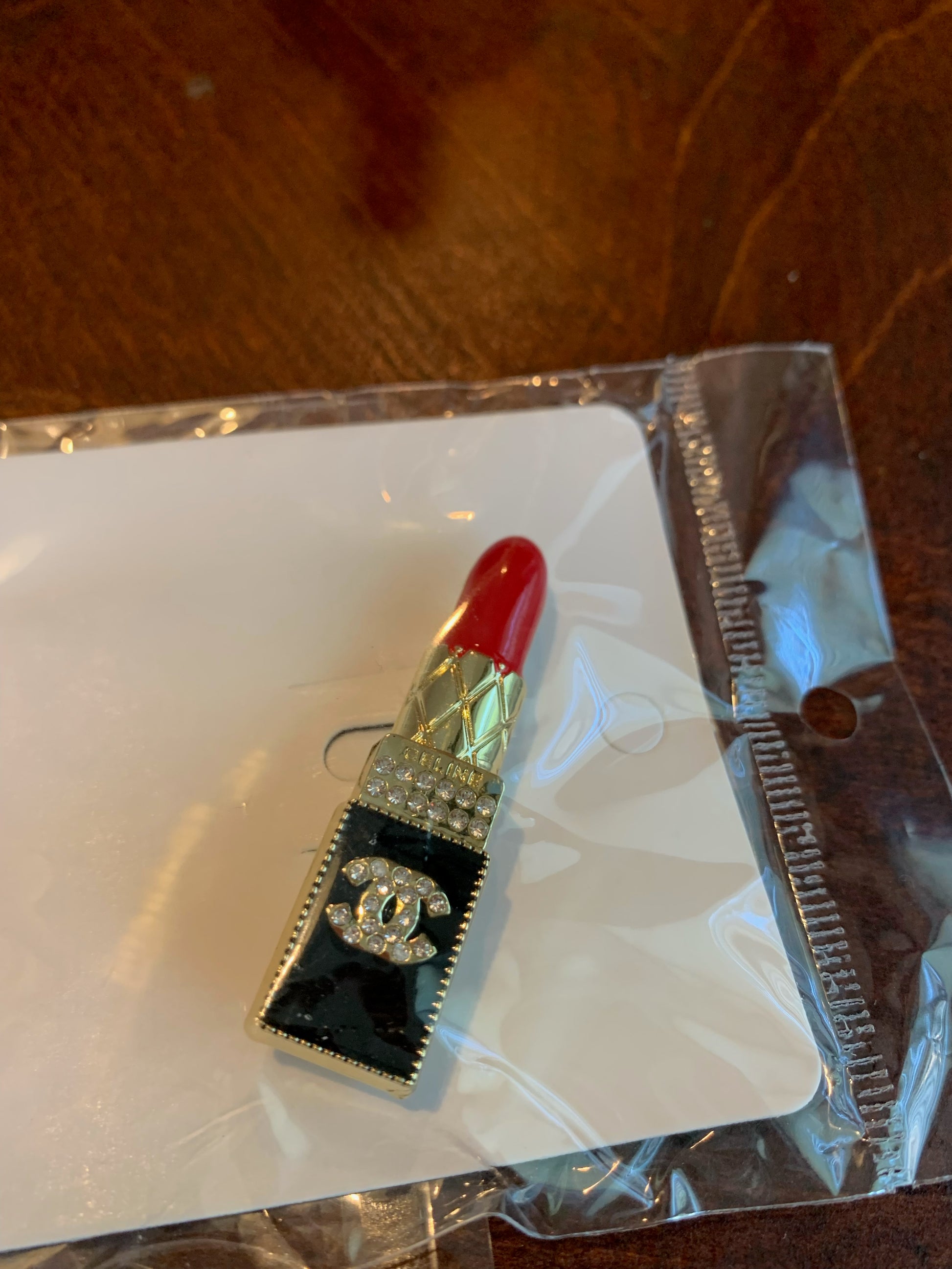 A Chanel-inspired lipstick-shaped brooch with a red lipstick tip, black and gold body, rhinestone details, and a CC logo, chanel inspired brooches